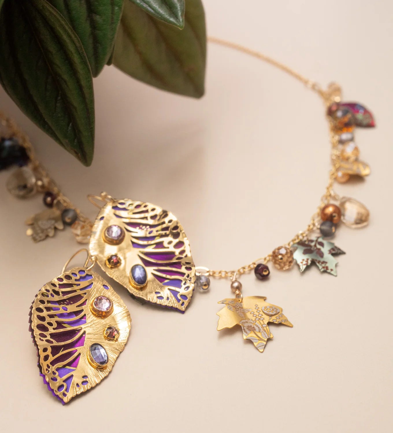 A gold necklace with leaf-shaped pendants, gemstone accents, and small charms rests on a beige surface beside green leaves, pairing beautifully with Holly Yashi's Perennial Elm Purple Earrings or other unique niobium jewelry.