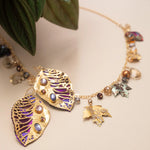 A gold necklace with leaf-shaped pendants, gemstone accents, and small charms rests on a beige surface beside green leaves, pairing beautifully with Holly Yashi's Perennial Elm Purple Earrings or other unique niobium jewelry.