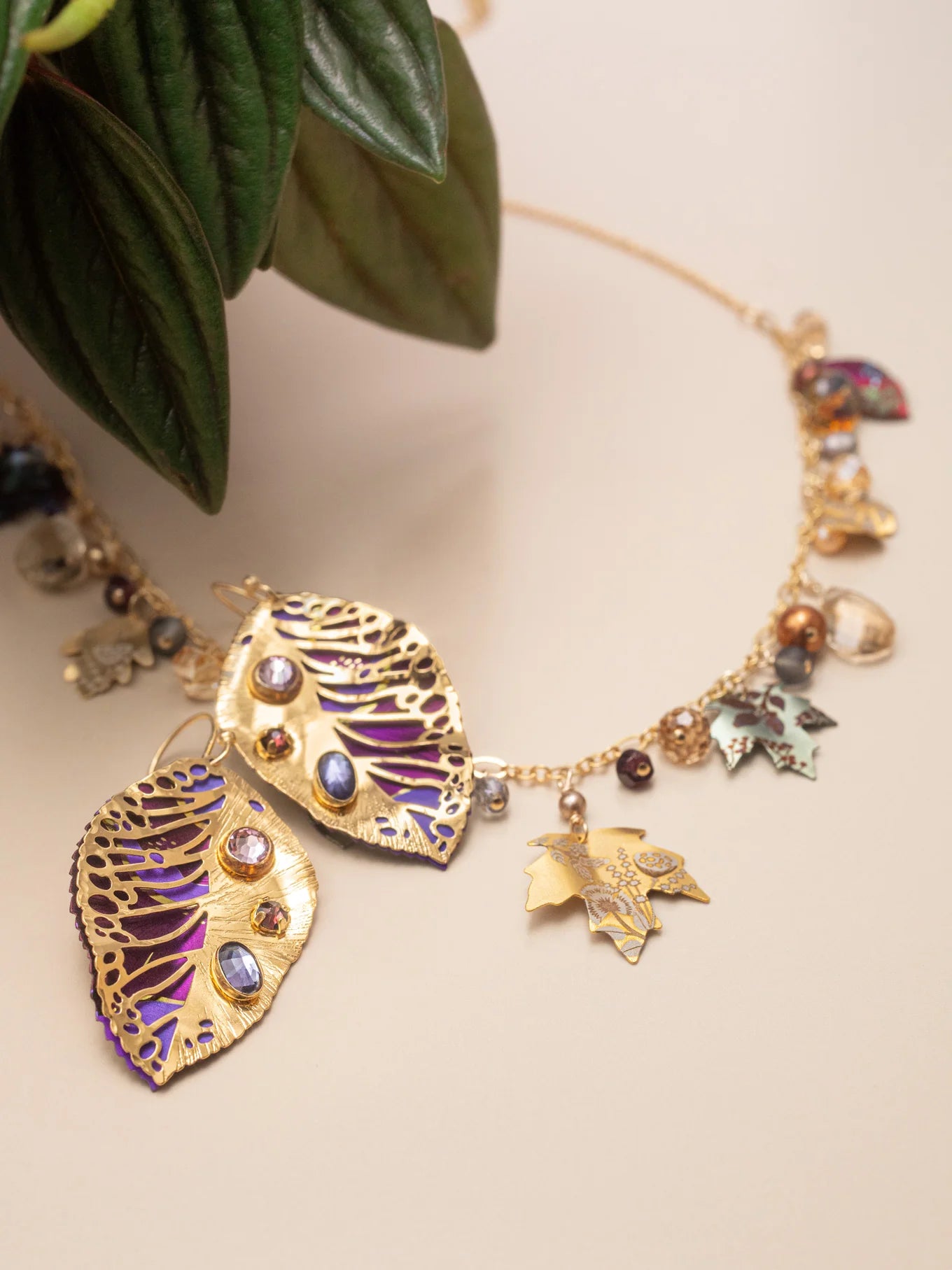 A gold necklace with leaf-shaped pendants, gemstone accents, and small charms rests on a beige surface beside green leaves, pairing beautifully with Holly Yashi's Perennial Elm Purple Earrings or other unique niobium jewelry.