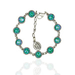 The 10 Simple Drops Teal Bracelet by Anne Koplik Designs, Inc. features round blue-green stones in silver iridescent beaded settings, linked with a lobster clasp and engraved charm reading "NOUS SOMMES TOUS DES HÉROS," shown on white background.