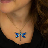 A woman in a black top wears the Anne Koplik Designs, Inc. Large Enamel/Stone Dragonfly Necklace—featuring a gold chain and blue hand-enameled pendant with crystal accents—her brown hair over her shoulder, smiling softly.