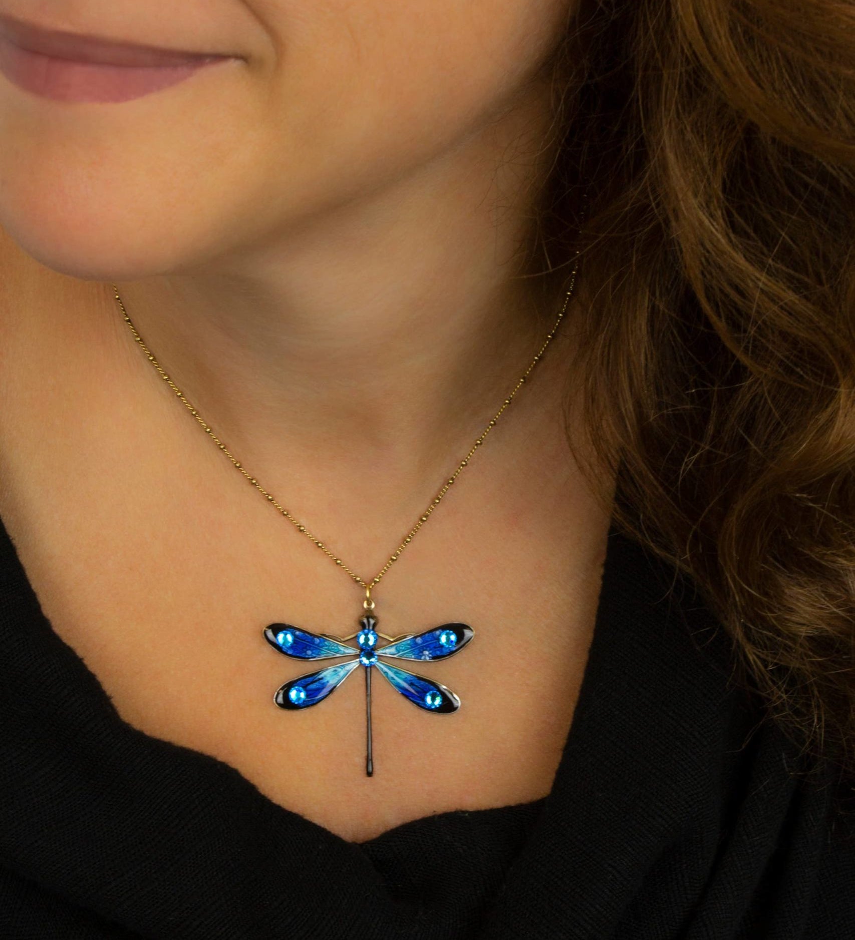 A woman in a black top wears the Anne Koplik Designs, Inc. Large Enamel/Stone Dragonfly Necklace—featuring a gold chain and blue hand-enameled pendant with crystal accents—her brown hair over her shoulder, smiling softly.