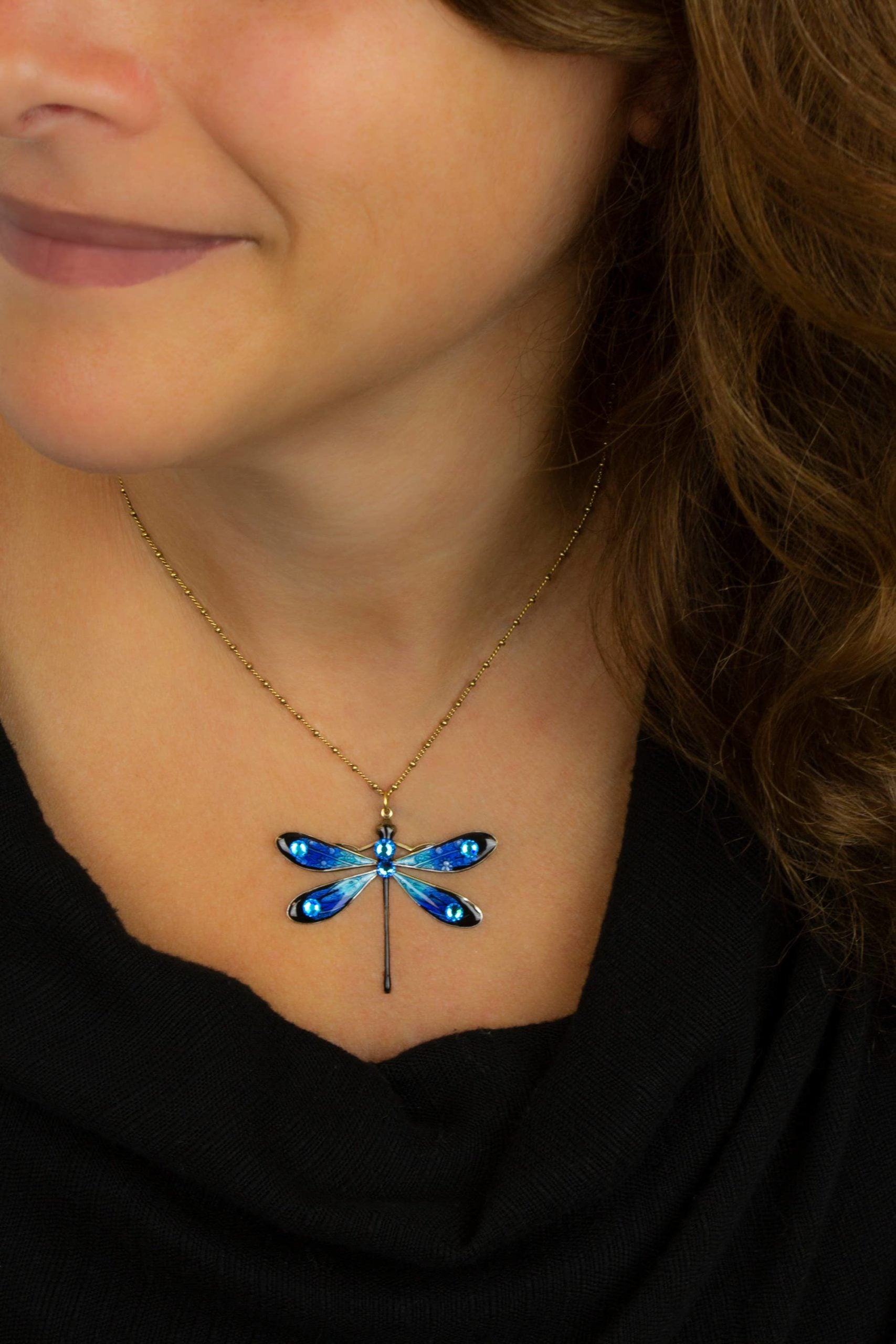 A woman in a black top wears the Anne Koplik Designs, Inc. Large Enamel/Stone Dragonfly Necklace—featuring a gold chain and blue hand-enameled pendant with crystal accents—her brown hair over her shoulder, smiling softly.
