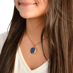 A woman with long brown hair in a white shirt wears the Anne Koplik Designs, Inc. 1/2 Moon Face/Stars Necklace, a gold enameled piece with blue and yellow constellation accents, highlighting elegant handcrafted jewelry.