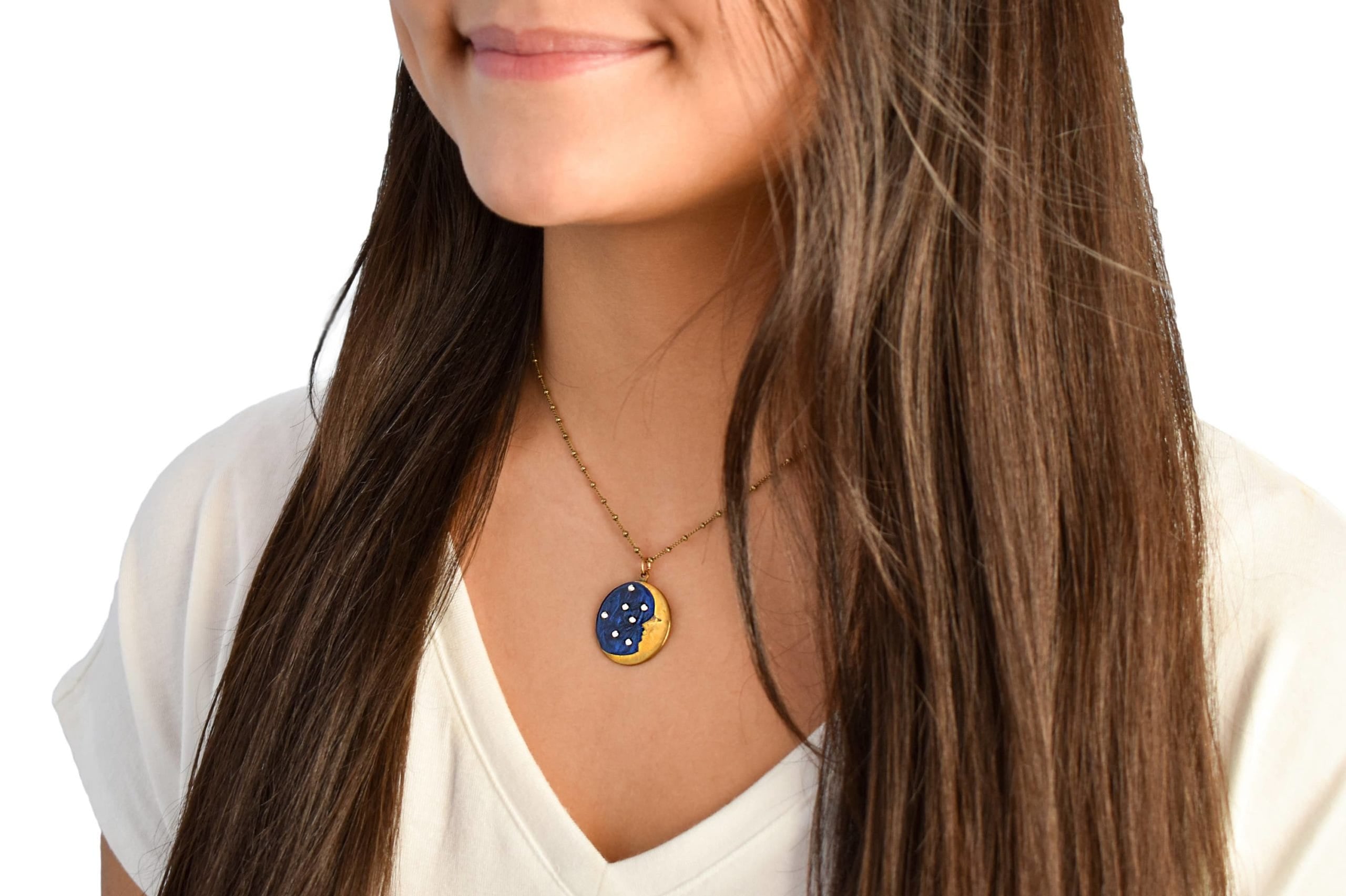 A woman with long brown hair in a white shirt wears the Anne Koplik Designs, Inc. 1/2 Moon Face/Stars Necklace, a gold enameled piece with blue and yellow constellation accents, highlighting elegant handcrafted jewelry.