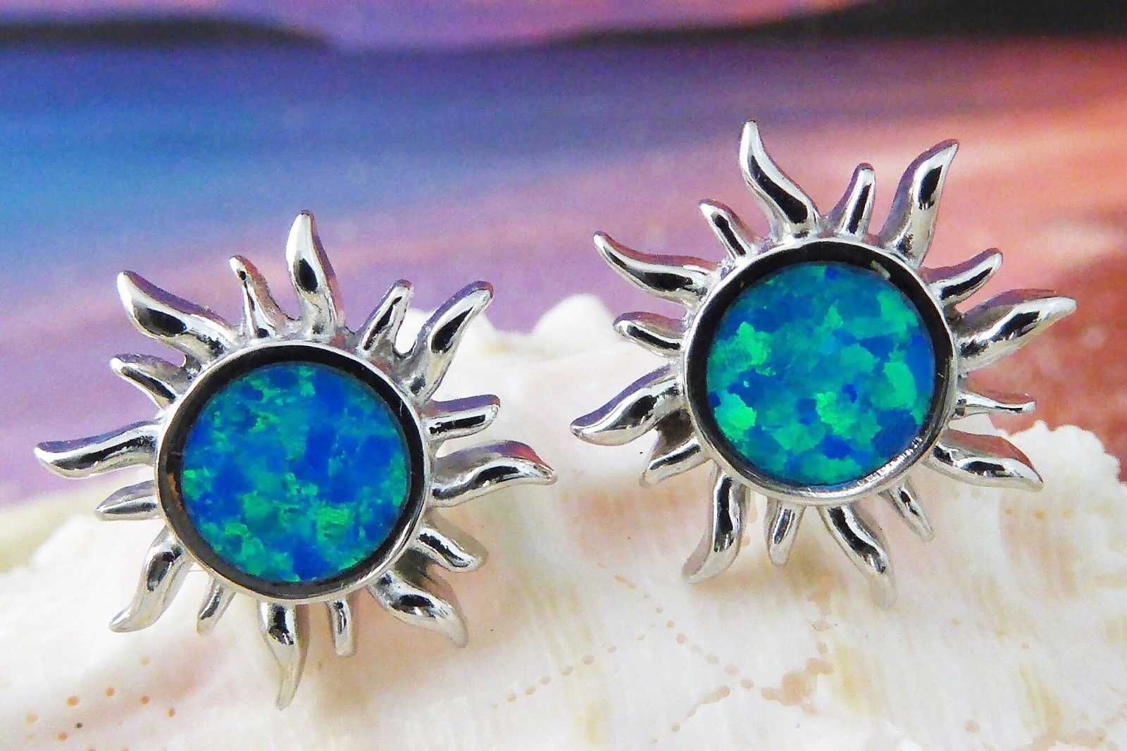 The Jewelry Network Caribbean Opal Sun Post Earrings feature sun-shaped rhodium plated 925 sterling silver with blue-green synthetic opal centers, displayed on a white seashell against a softly blurred gradient background.