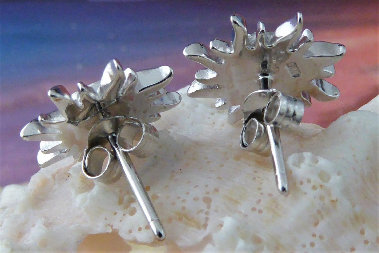 The Caribbean Opal Sun Post Earrings by Jewelry Network, crafted in rhodium plated 925 Sterling Silver with a sunburst design, are displayed on a pale seashell against a softly blurred background in purple, blue, and pink hues.