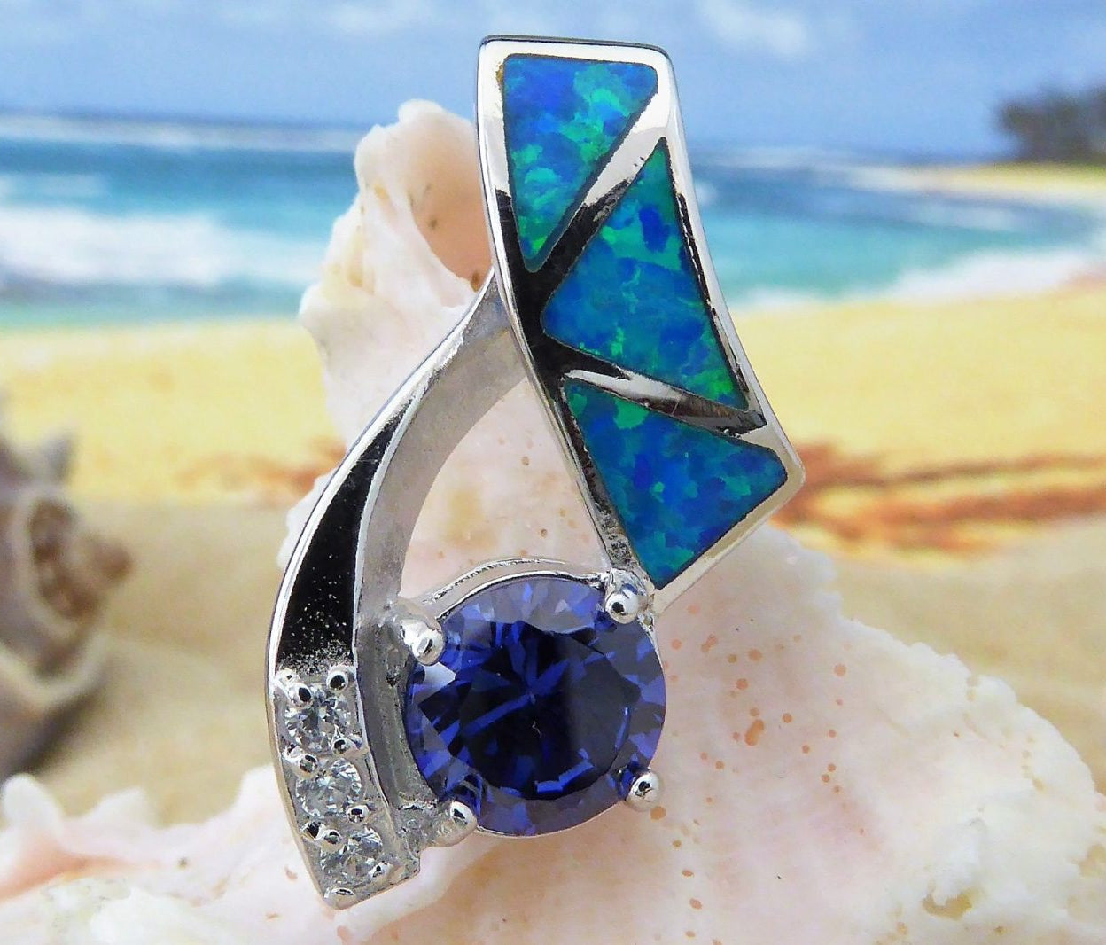 The Jewelry Network Caribbean Opal 3 Triangles/Round Amethyst pendant (pendant only) is displayed on a seashell, with turquoise geometric inlays and a round amethyst against a blurred beach and ocean background.