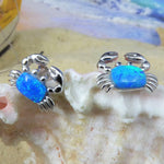 Two Jewelry Network Caribbean Opal Crab Post Earrings, adorned with vibrant blue opal stones, are displayed on a seashell with a softly blurred sandy beach and ocean in the background.