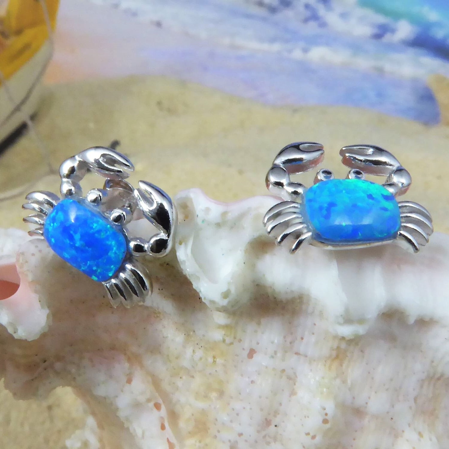 Two Jewelry Network Caribbean Opal Crab Post Earrings, adorned with vibrant blue opal stones, are displayed on a seashell with a softly blurred sandy beach and ocean in the background.