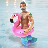 A bearded merman with a tattoo stands waist-deep in water, sporting a pink Flaming Oh! Ornament float. His shiny turquoise tail sparkles like holiday decor against the calm, reflective water.