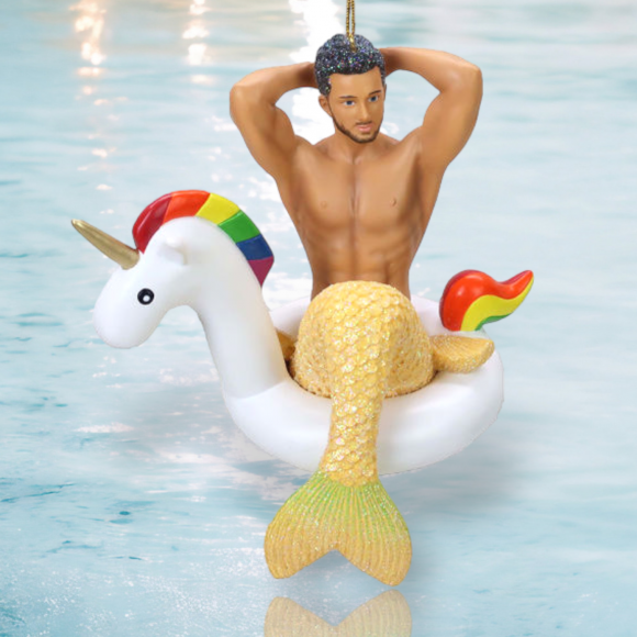 The Mr. Unicorn Ornament by December Diamonds features a hand-painted merman with a glittery tail lounging in a unicorn float, complete with a rainbow mane, horn, and heart decoration. The soft blue water enhances the summery charm of this holiday decor piece.