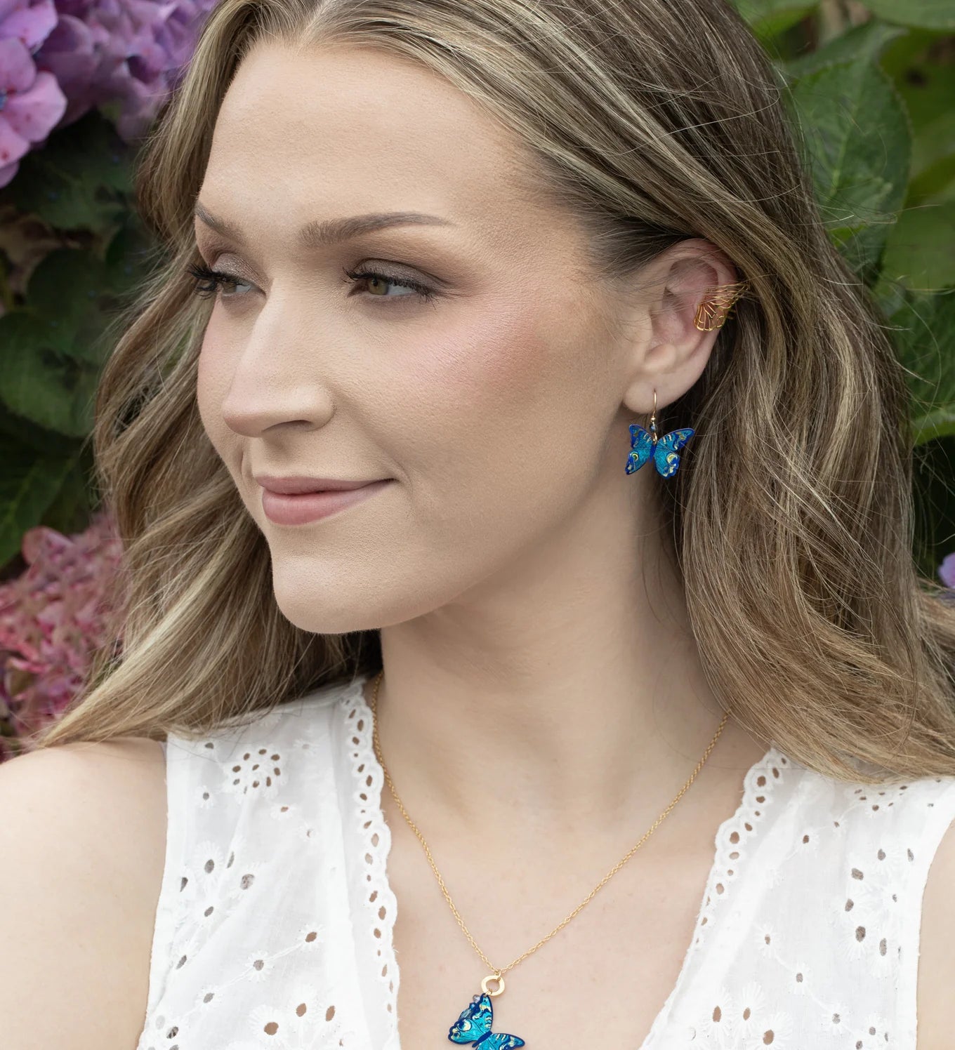 A woman with wavy blonde hair wears a white sleeveless top, blue butterfly earrings, and the Holly Yashi NK: Mariposa Butterfly Charm Pendant Blue Radiance necklace. She smiles outside with purple hydrangeas behind her.