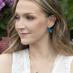 A woman with wavy blonde hair wears a white sleeveless top, blue butterfly earrings, and the Holly Yashi NK: Mariposa Butterfly Charm Pendant Blue Radiance necklace. She smiles outside with purple hydrangeas behind her.