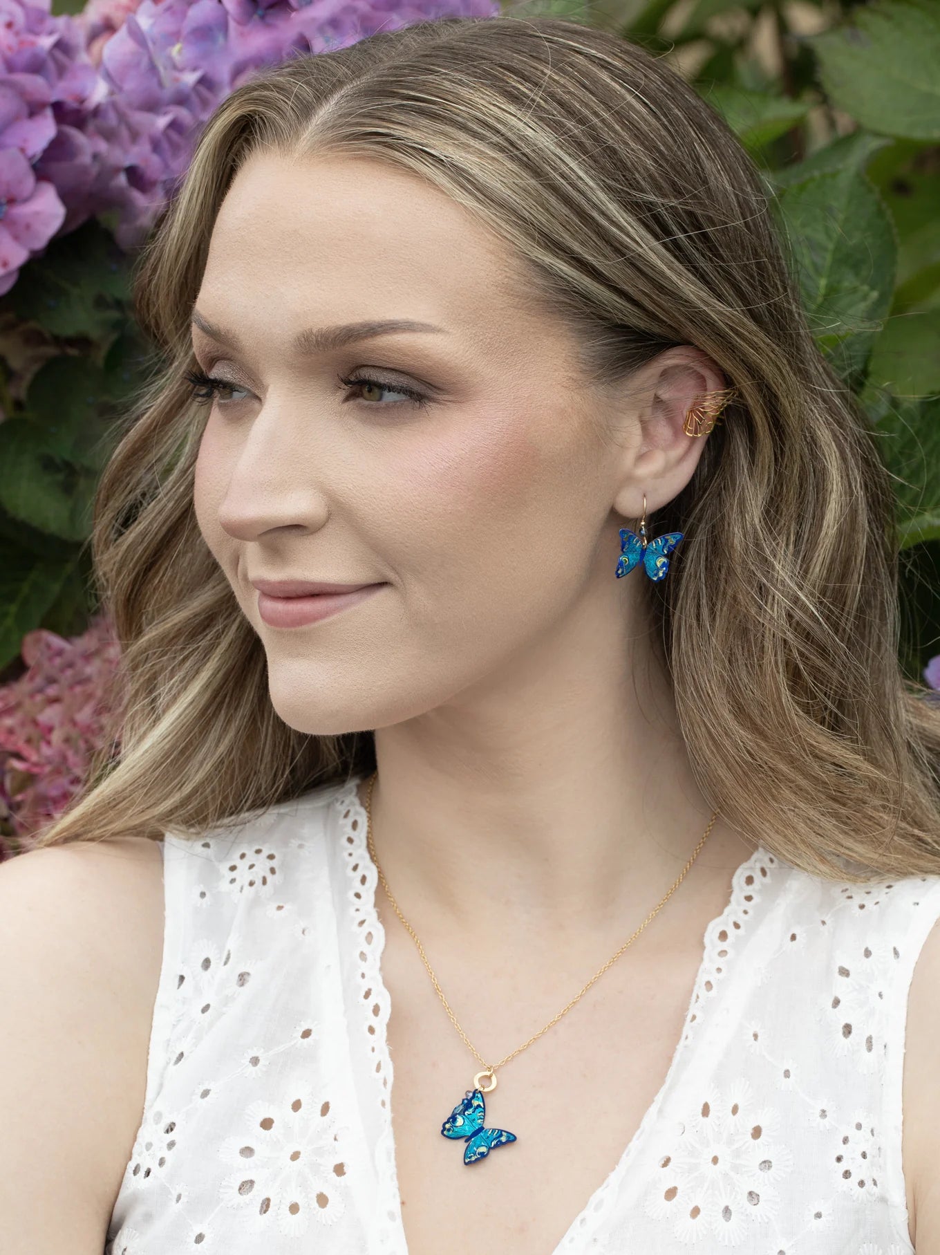 A woman with wavy blonde hair wears a white sleeveless top, blue butterfly earrings, and the Holly Yashi NK: Mariposa Butterfly Charm Pendant Blue Radiance necklace. She smiles outside with purple hydrangeas behind her.