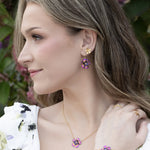 A woman with wavy blonde hair wears a floral white dress and Holly Yashi’s Blissful Bloom Bonnie Blue Earrings—hypoallergenic earrings featuring blue flower designs with sparkling crystal centers. Pink flowers and green leaves fill the background.