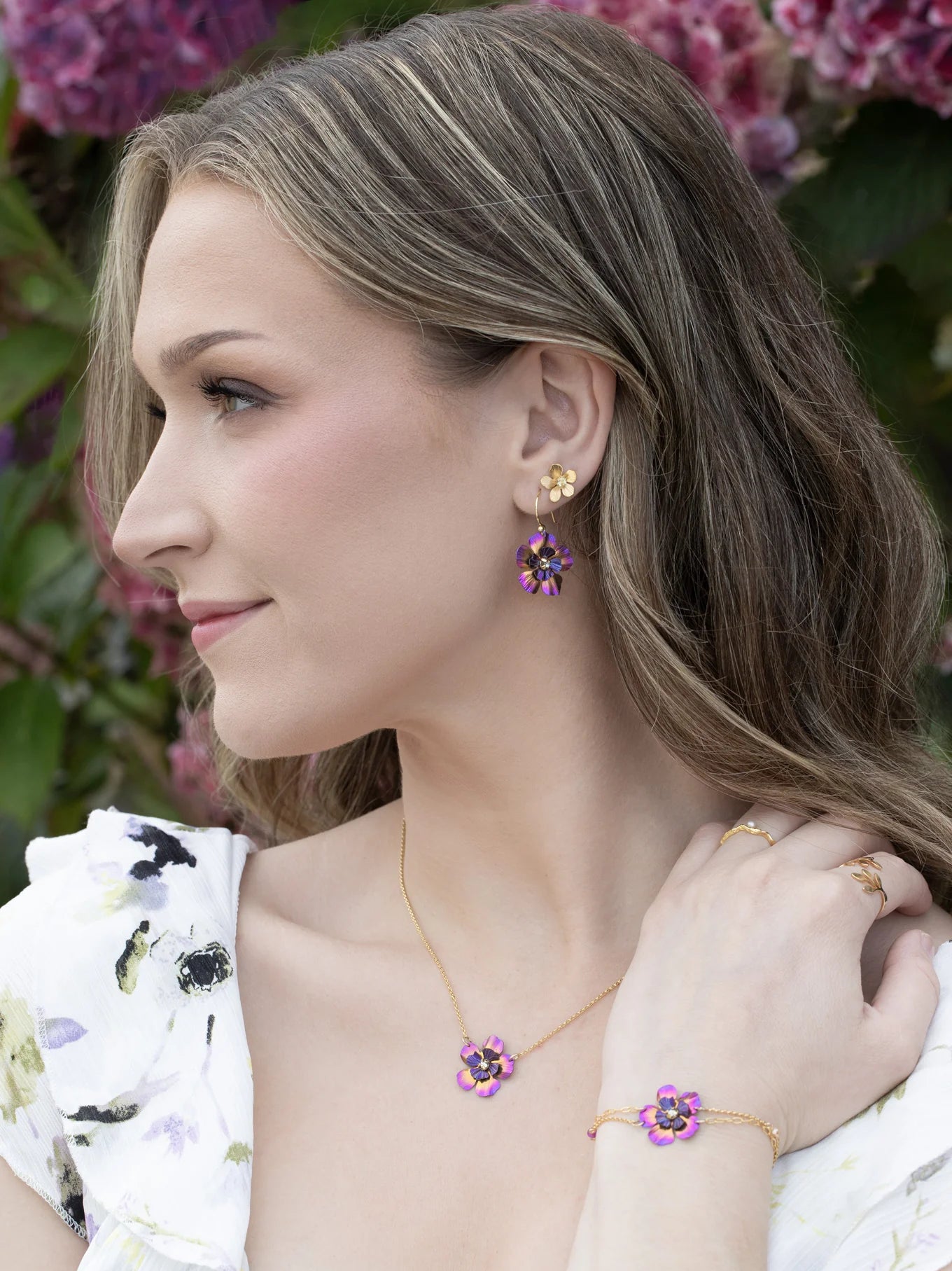 A woman with wavy blonde hair wears a floral white dress and Holly Yashi’s Blissful Bloom Bonnie Blue Earrings—hypoallergenic earrings featuring blue flower designs with sparkling crystal centers. Pink flowers and green leaves fill the background.