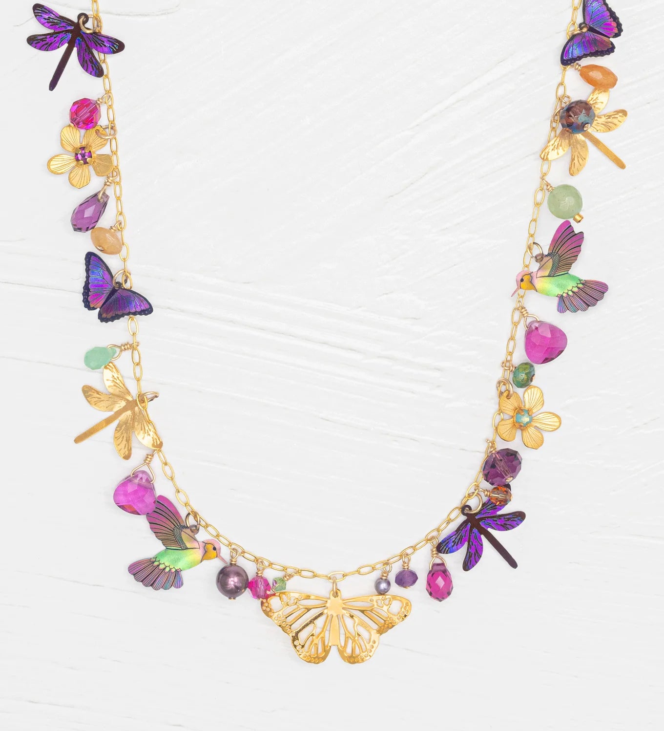 The Garden Party Gold Charms Necklace by Holly Yashi features a gold chain embellished with colorful niobium butterflies, dragonflies, flowers, and gemstone charms. Shown on a white textured background, this unique necklace offers an adjustable chain for the perfect fit.