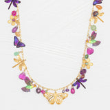 The Garden Party Gold Charms Necklace by Holly Yashi features a gold chain embellished with colorful niobium butterflies, dragonflies, flowers, and gemstone charms. Shown on a white textured background, this unique necklace offers an adjustable chain for the perfect fit.