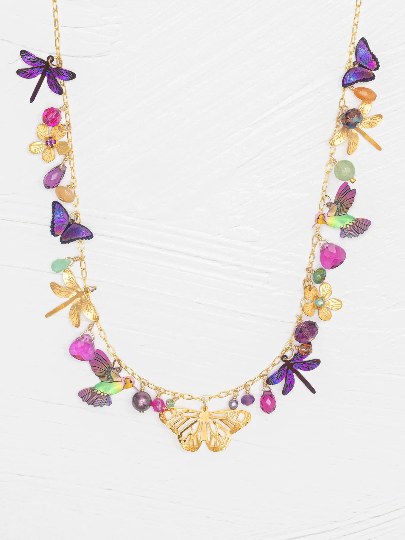 The Garden Party Gold Charms Necklace by Holly Yashi features a gold chain embellished with colorful niobium butterflies, dragonflies, flowers, and gemstone charms. Shown on a white textured background, this unique necklace offers an adjustable chain for the perfect fit.