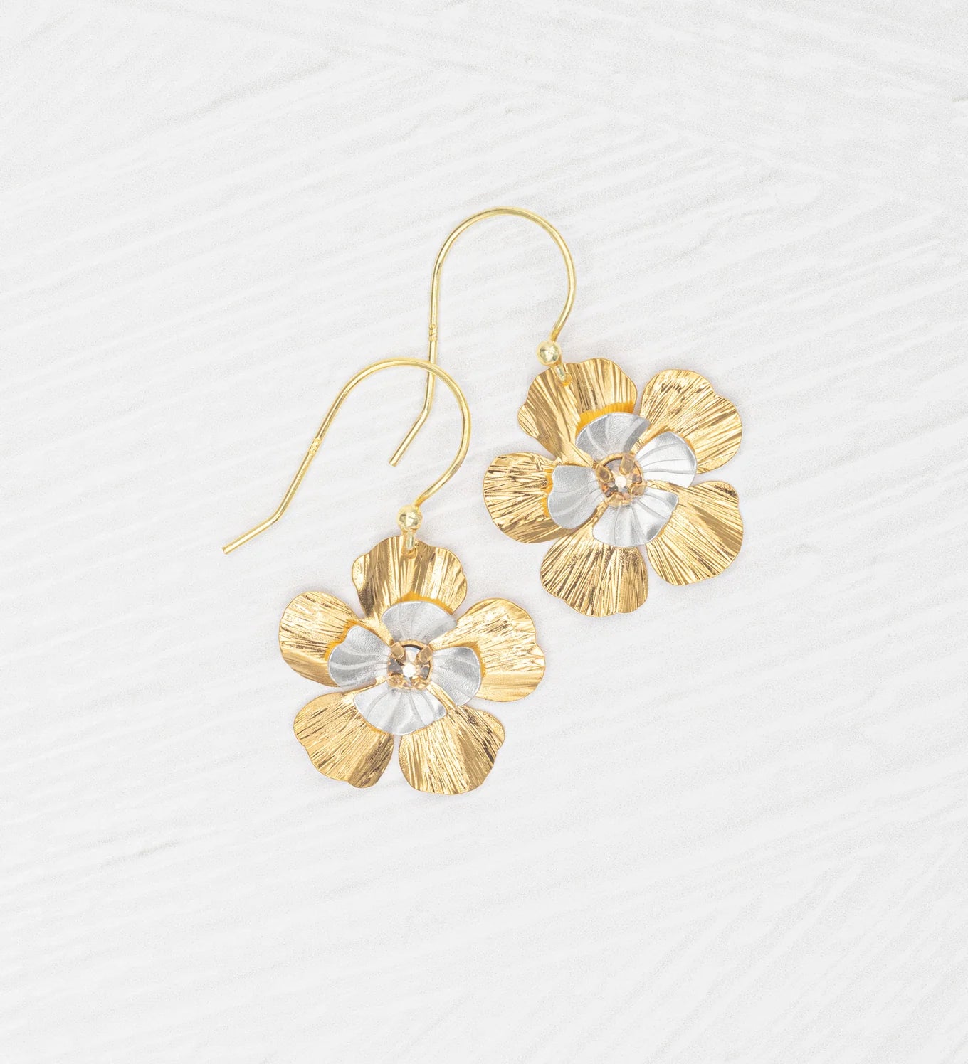 The Blissful Bloom Gold & Silver Earrings by Holly Yashi are hypoallergenic, flower-shaped dangles with textured gold and silver petals and a central gem, shown on a white textured surface.