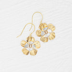 The Blissful Bloom Gold & Silver Earrings by Holly Yashi are hypoallergenic, flower-shaped dangles with textured gold and silver petals and a central gem, shown on a white textured surface.