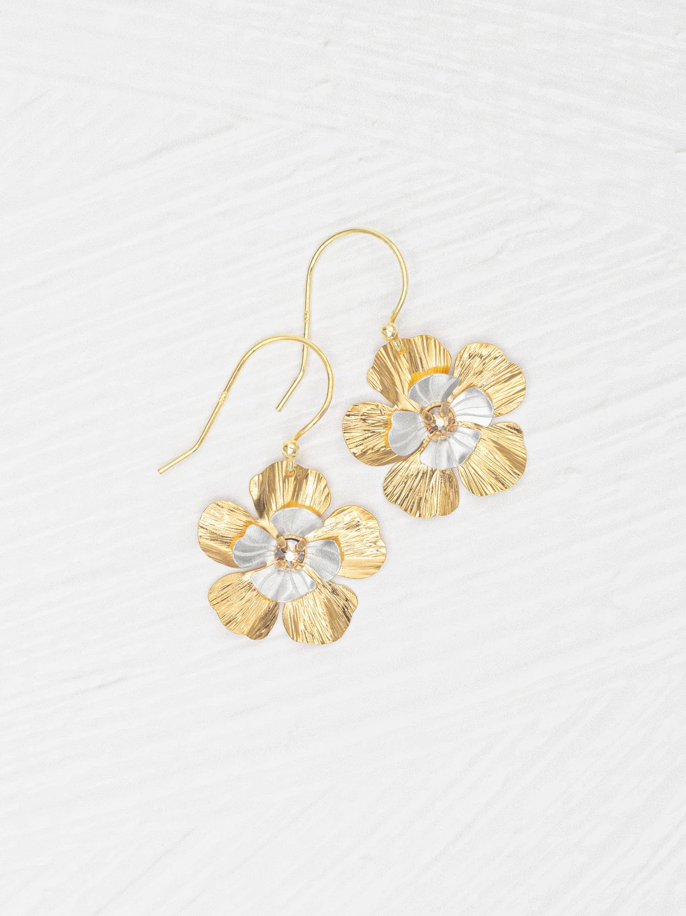 The Blissful Bloom Gold & Silver Earrings by Holly Yashi are hypoallergenic, flower-shaped dangles with textured gold and silver petals and a central gem, shown on a white textured surface.