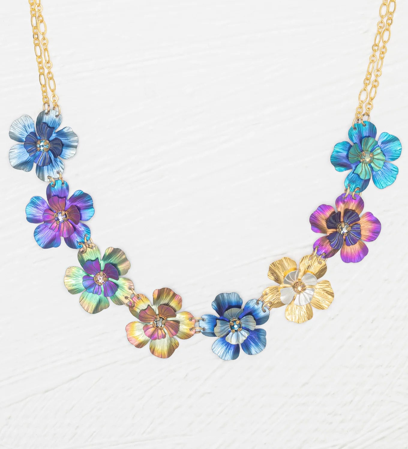 The Holly Yashi Blissful Bouquet Multicolor Necklace showcases a gold chain with eight handcrafted niobium flower pendants in blue, purple, gold, and multicolor finishes, each featuring crystal accents and arranged symmetrically.