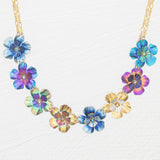 The Holly Yashi Blissful Bouquet Multicolor Necklace showcases a gold chain with eight handcrafted niobium flower pendants in blue, purple, gold, and multicolor finishes, each featuring crystal accents and arranged symmetrically.