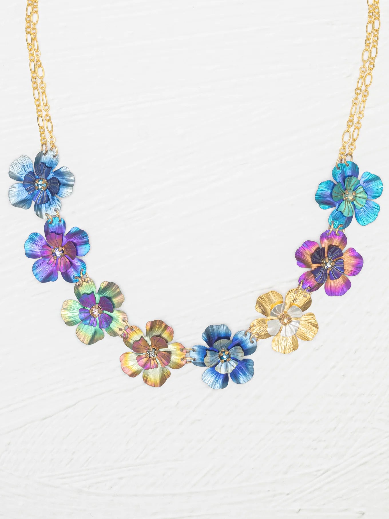 The Holly Yashi Blissful Bouquet Multicolor Necklace showcases a gold chain with eight handcrafted niobium flower pendants in blue, purple, gold, and multicolor finishes, each featuring crystal accents and arranged symmetrically.