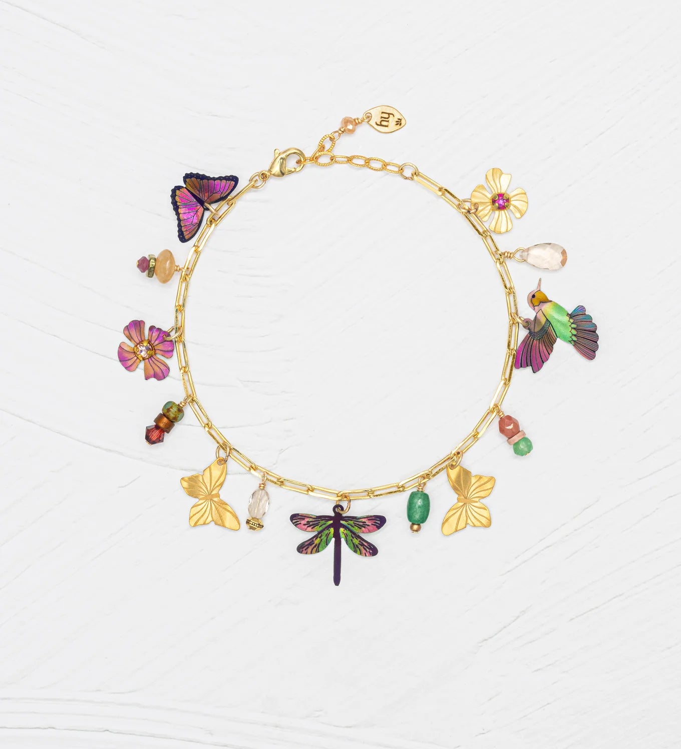The Holly Yashi Garden Whimsy Gold Charms Bracelet features colorful charms—including butterflies, flowers, a dragonfly, and various beads—displayed on a white textured background.