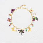 The Holly Yashi Garden Whimsy Gold Charms Bracelet features colorful charms—including butterflies, flowers, a dragonfly, and various beads—displayed on a white textured background.