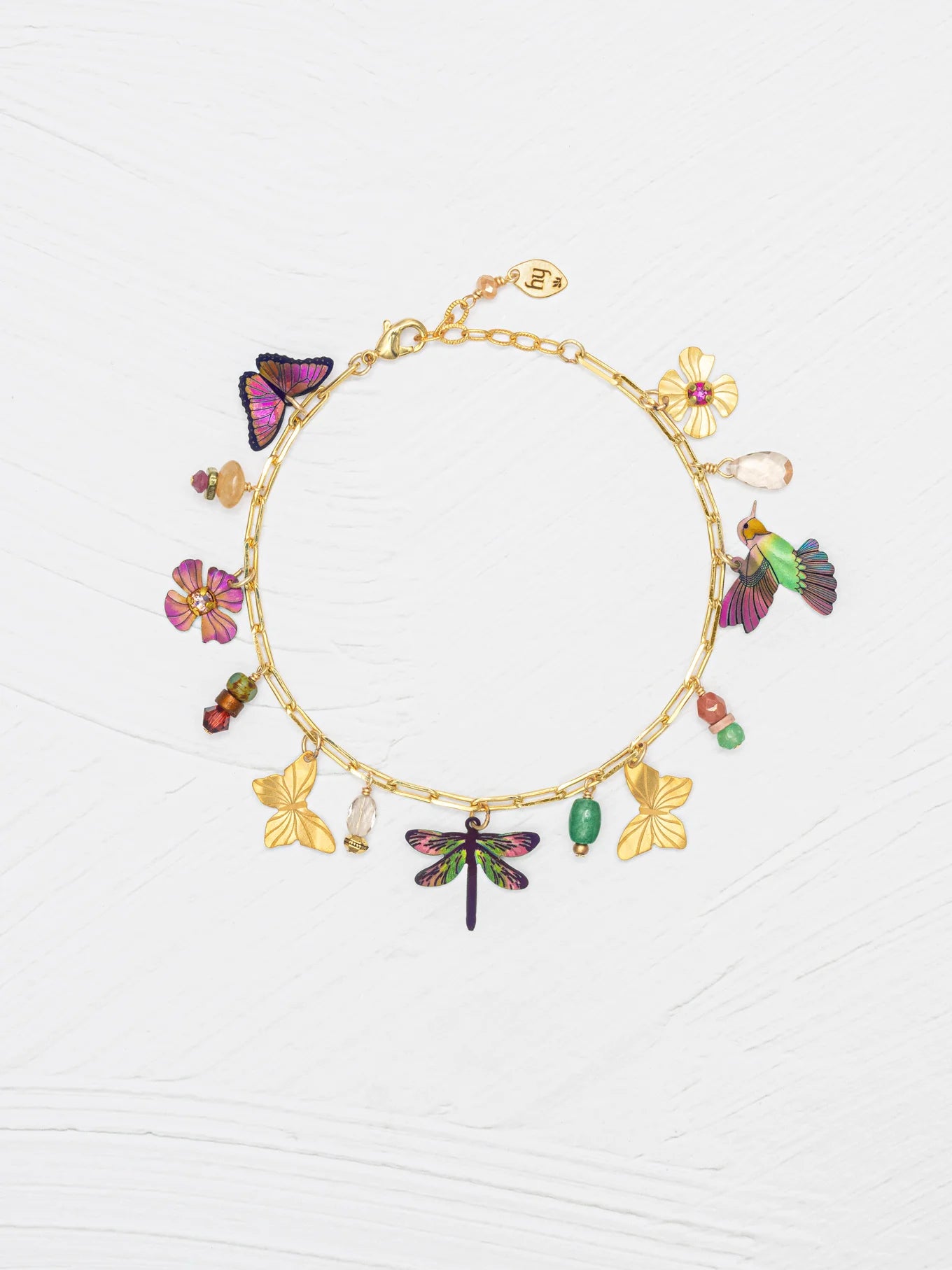 The Holly Yashi Garden Whimsy Gold Charms Bracelet features colorful charms—including butterflies, flowers, a dragonfly, and various beads—displayed on a white textured background.