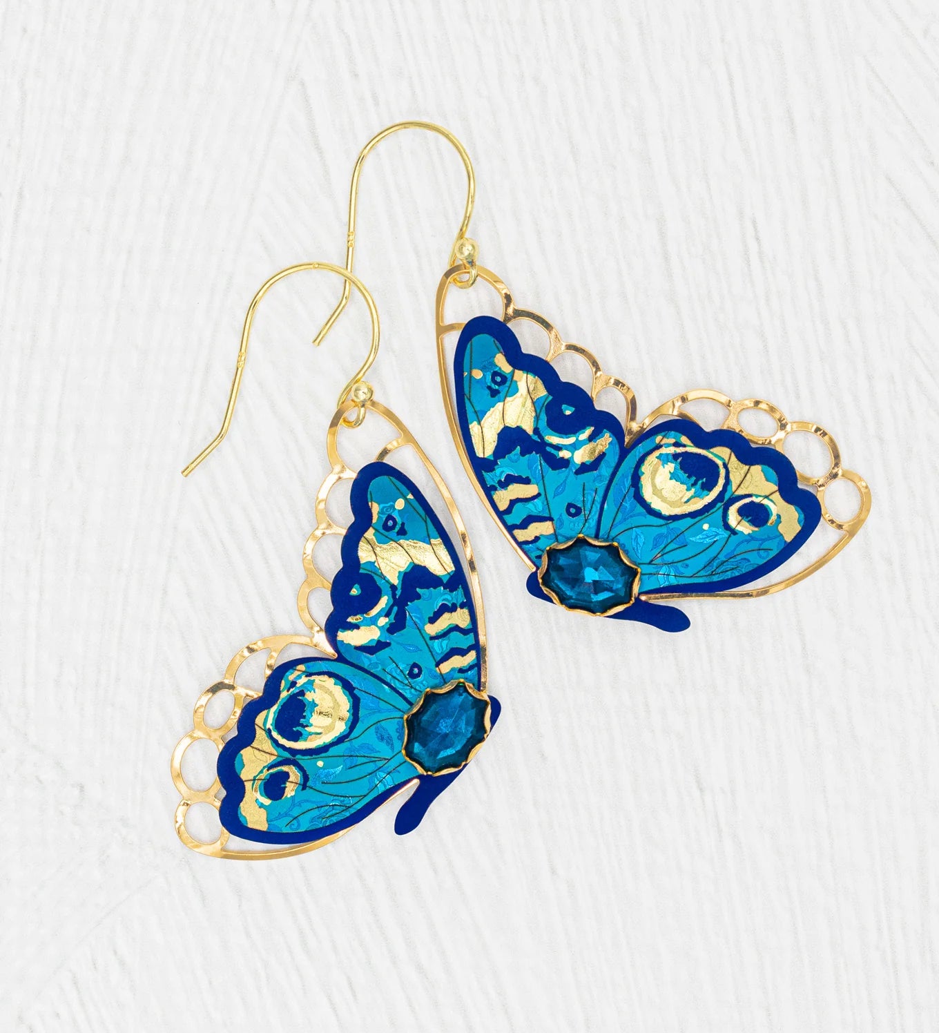 The Holly Yashi Mariposa Butterfly Wing Blue Radiance Earrings are gold-filled, hypoallergenic earrings with intricate blue butterfly designs and gold detailing, shown on a textured white background.