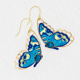 The Holly Yashi Mariposa Butterfly Wing Blue Radiance Earrings are gold-filled, hypoallergenic earrings with intricate blue butterfly designs and gold detailing, shown on a textured white background.