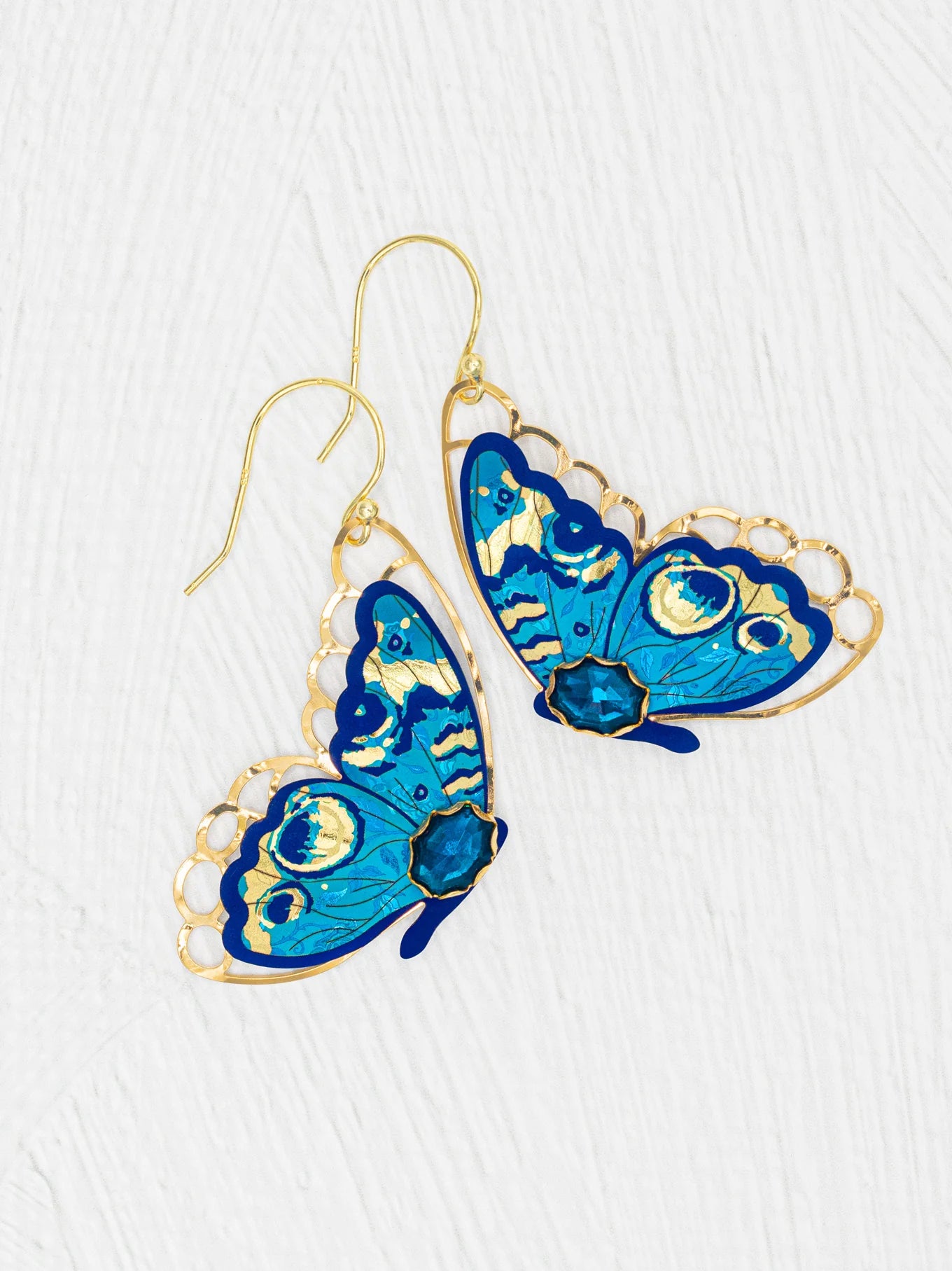 The Holly Yashi Mariposa Butterfly Wing Blue Radiance Earrings are gold-filled, hypoallergenic earrings with intricate blue butterfly designs and gold detailing, shown on a textured white background.