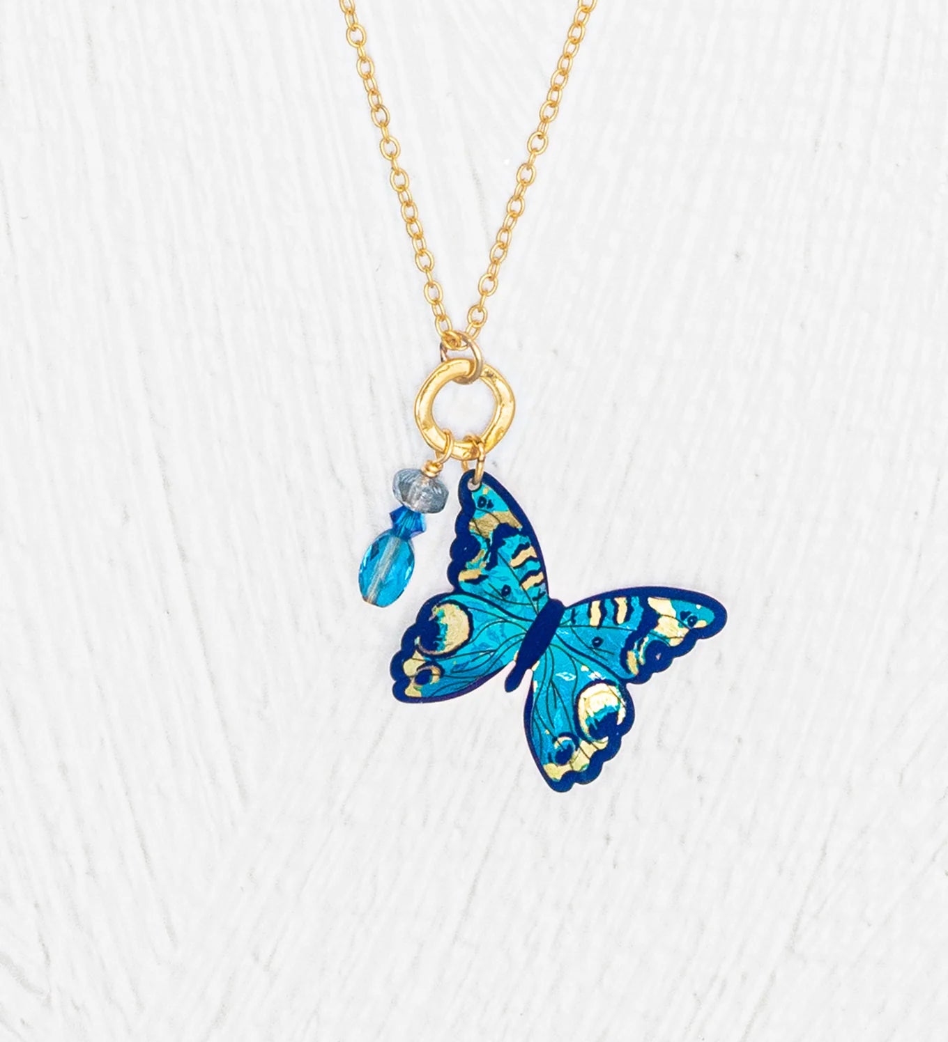 The Holly Yashi NK: Mariposa Butterfly Charm Pendant Blue Radiance Necklace features a gold adjustable chain with a blue butterfly charm and two small blue bead charms, set against a white textured background. Ideal for elegant jewelry lovers.