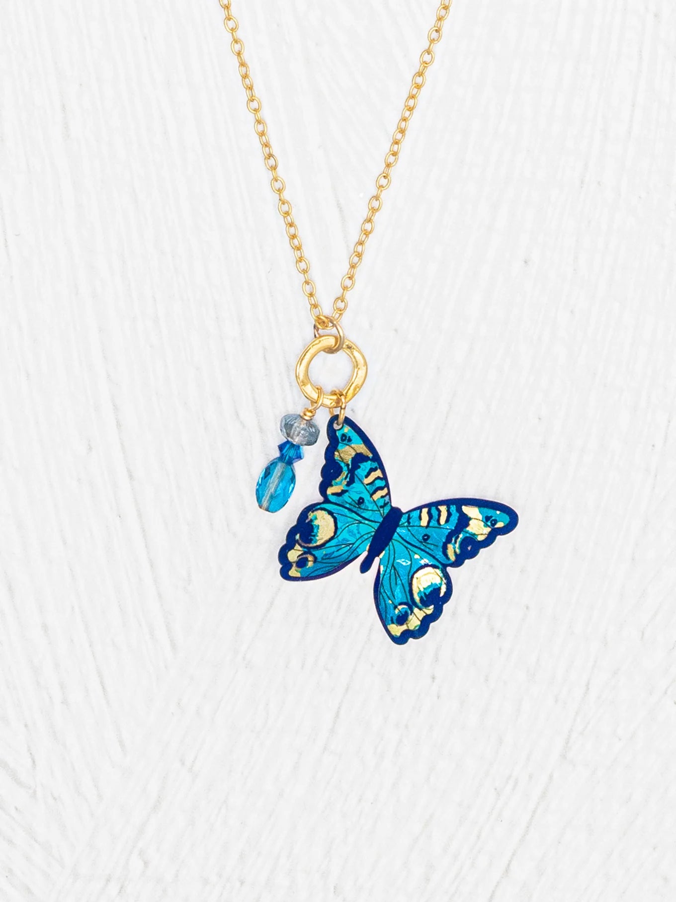 The Holly Yashi NK: Mariposa Butterfly Charm Pendant Blue Radiance Necklace features a gold adjustable chain with a blue butterfly charm and two small blue bead charms, set against a white textured background. Ideal for elegant jewelry lovers.
