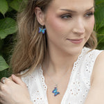 A woman in a white eyelet top wears Holly Yashi's Mariposa Butterfly Blue Radiance earrings, paired with matching gold-filled butterfly necklace, hypoallergenic silver bracelet and ring, her hair down as she smiles softly near green foliage.