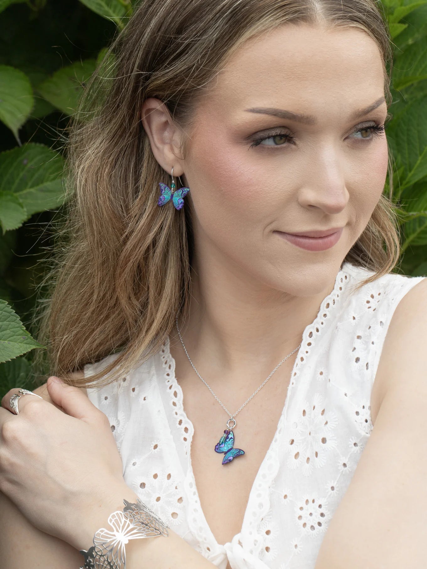 A woman in a white eyelet top wears Holly Yashi's Mariposa Butterfly Blue Radiance earrings, paired with matching gold-filled butterfly necklace, hypoallergenic silver bracelet and ring, her hair down as she smiles softly near green foliage.