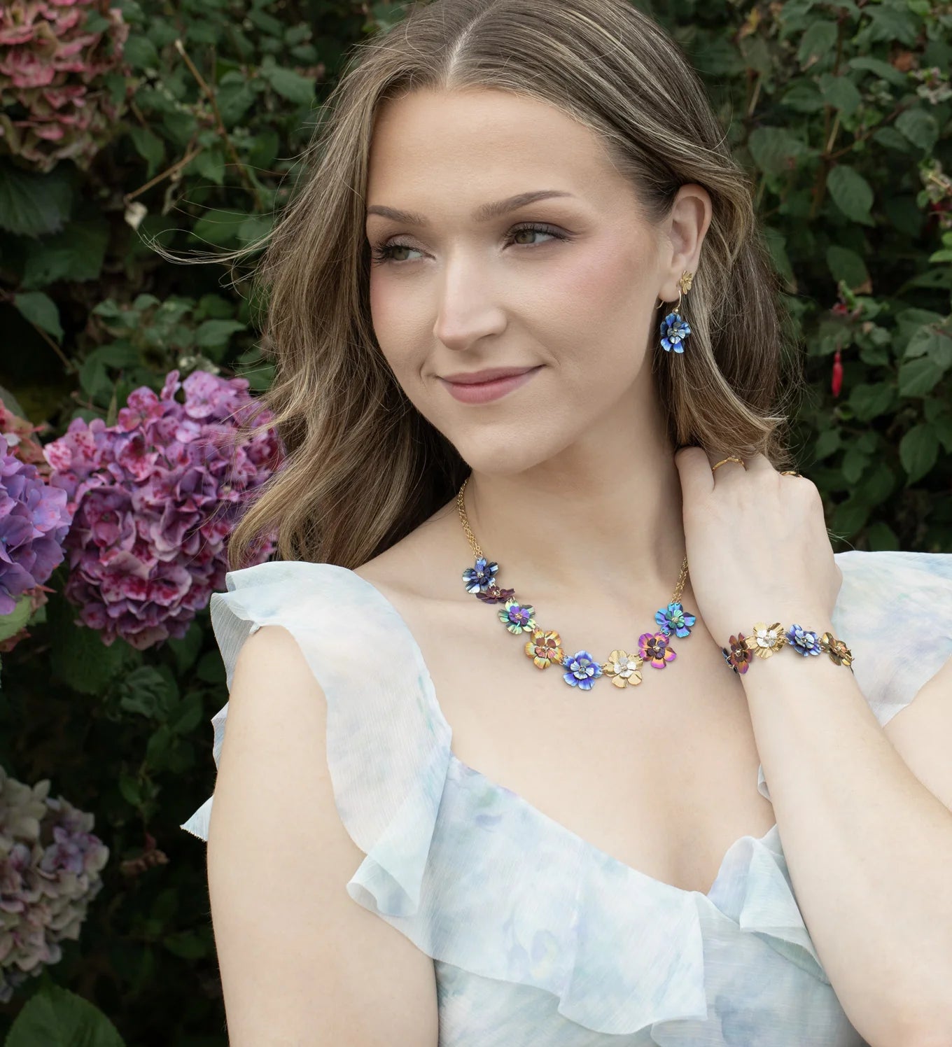 A woman with wavy hair, outdoors near hydrangeas, wears a light ruffled dress and the Holly Yashi Blissful Bouquet Multicolor Necklace with matching earrings and bracelet, gazing softly to the side.