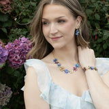 A woman with wavy hair, outdoors near hydrangeas, wears a light ruffled dress and the Holly Yashi Blissful Bouquet Multicolor Necklace with matching earrings and bracelet, gazing softly to the side.