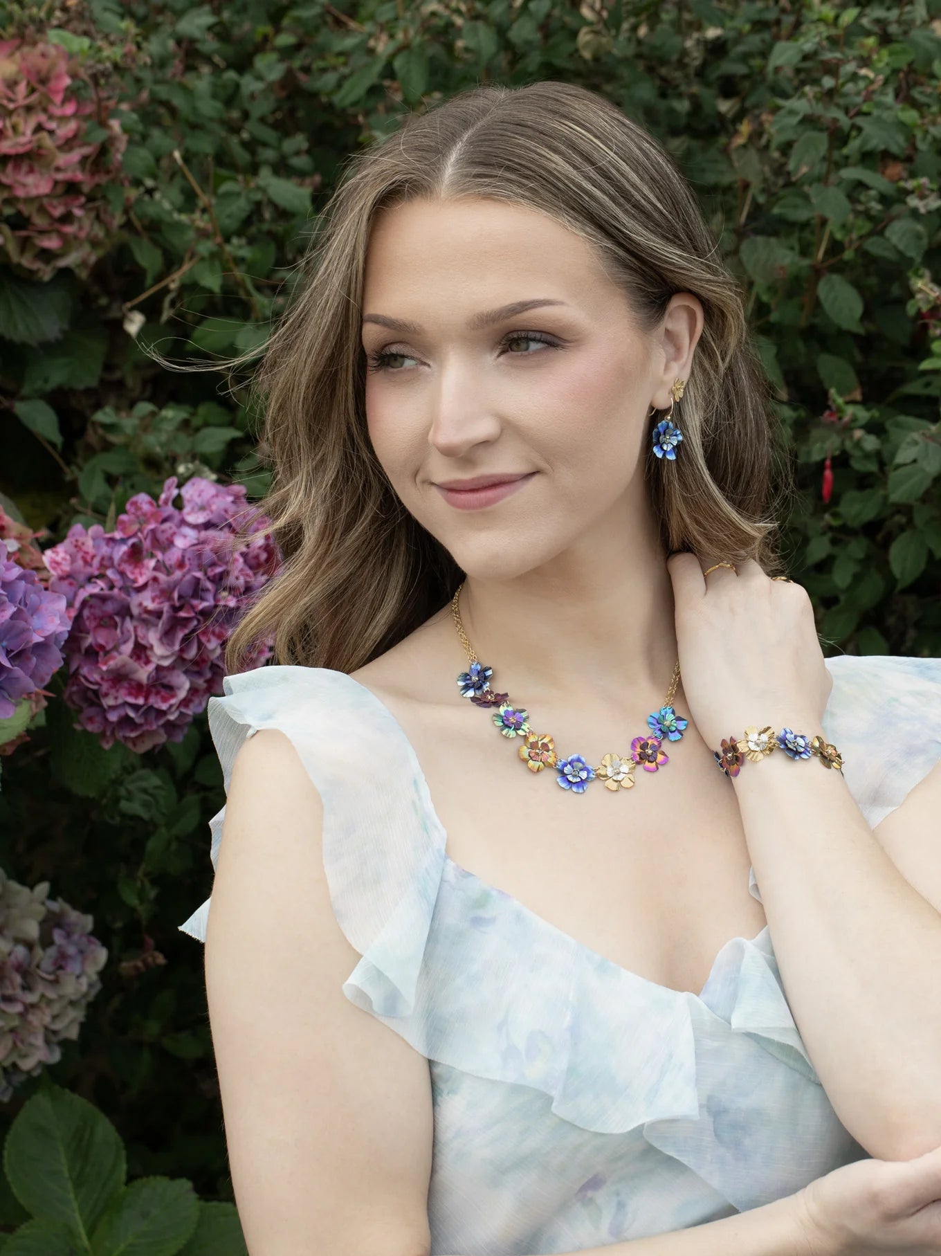 A woman with wavy hair, outdoors near hydrangeas, wears a light ruffled dress and the Holly Yashi Blissful Bouquet Multicolor Necklace with matching earrings and bracelet, gazing softly to the side.