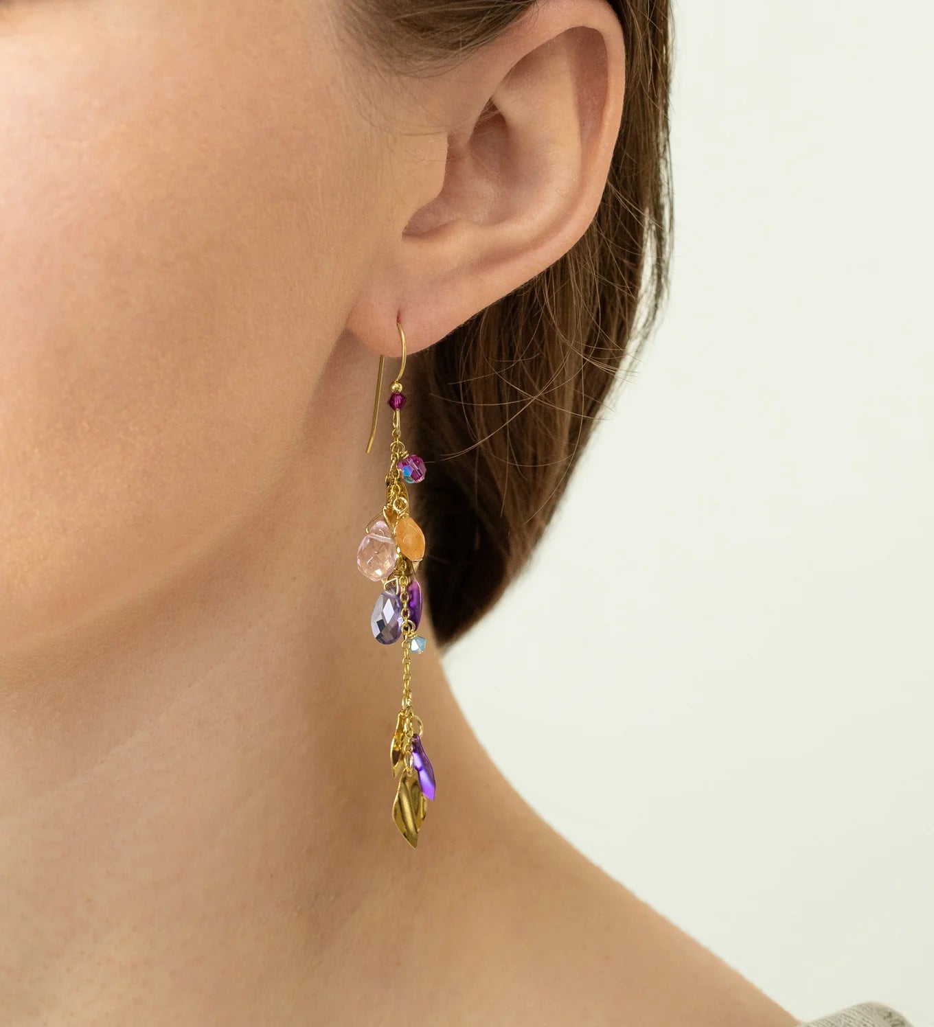 Close-up of a person wearing Holly Yashi Calliope Tropic Punch Earrings, featuring gold leaf, colorful beads, and charms. The hypoallergenic earrings shine as their hair is tucked behind the ear with a light-colored top.