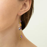 Close-up of a person wearing Holly Yashi Calliope Tropic Punch Earrings, featuring gold leaf, colorful beads, and charms. The hypoallergenic earrings shine as their hair is tucked behind the ear with a light-colored top.