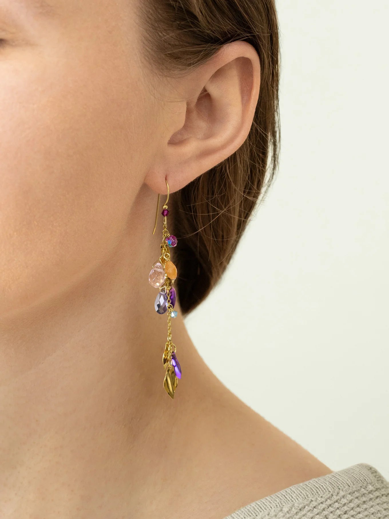 Close-up of a person wearing Holly Yashi Calliope Tropic Punch Earrings, featuring gold leaf, colorful beads, and charms. The hypoallergenic earrings shine as their hair is tucked behind the ear with a light-colored top.