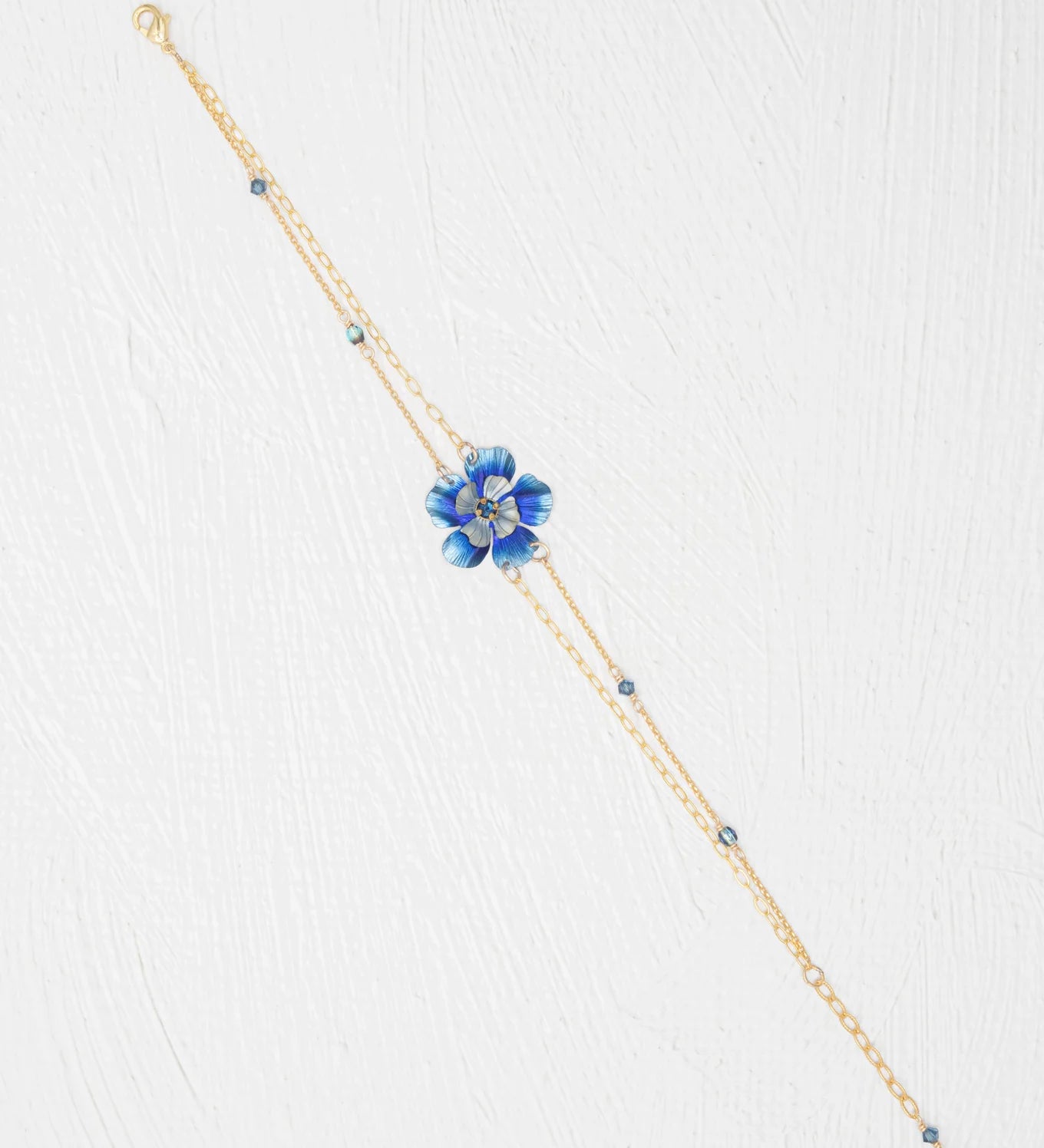 The Holly Yashi Blissful Bloom Double Strand Bonnie Blue Bracelet features a gold double strand with a blue flower charm, crystal center, and blue beads on hypoallergenic materials, shown on a white textured background.