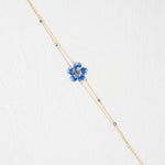 The Holly Yashi Blissful Bloom Double Strand Bonnie Blue Bracelet features a gold double strand with a blue flower charm, crystal center, and blue beads on hypoallergenic materials, shown on a white textured background.