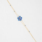 The Holly Yashi Blissful Bloom Double Strand Bonnie Blue Bracelet features a gold double strand with a blue flower charm, crystal center, and blue beads on hypoallergenic materials, shown on a white textured background.