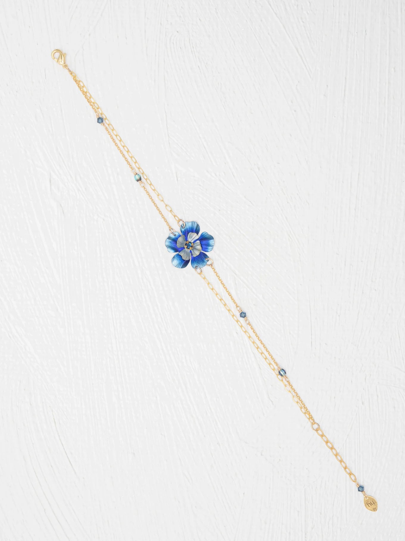 The Holly Yashi Blissful Bloom Double Strand Bonnie Blue Bracelet features a gold double strand with a blue flower charm, crystal center, and blue beads on hypoallergenic materials, shown on a white textured background.